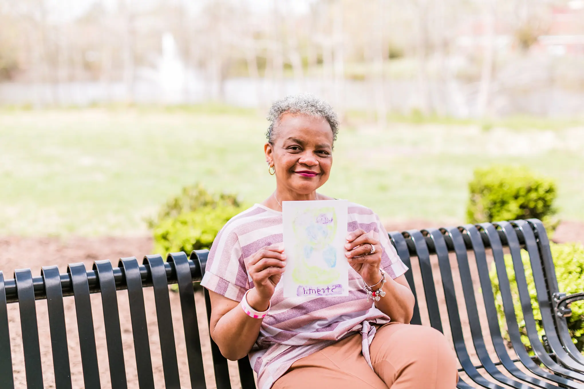 An elderly Black woman is sitting on a bench outside and holding up a self-made painting.