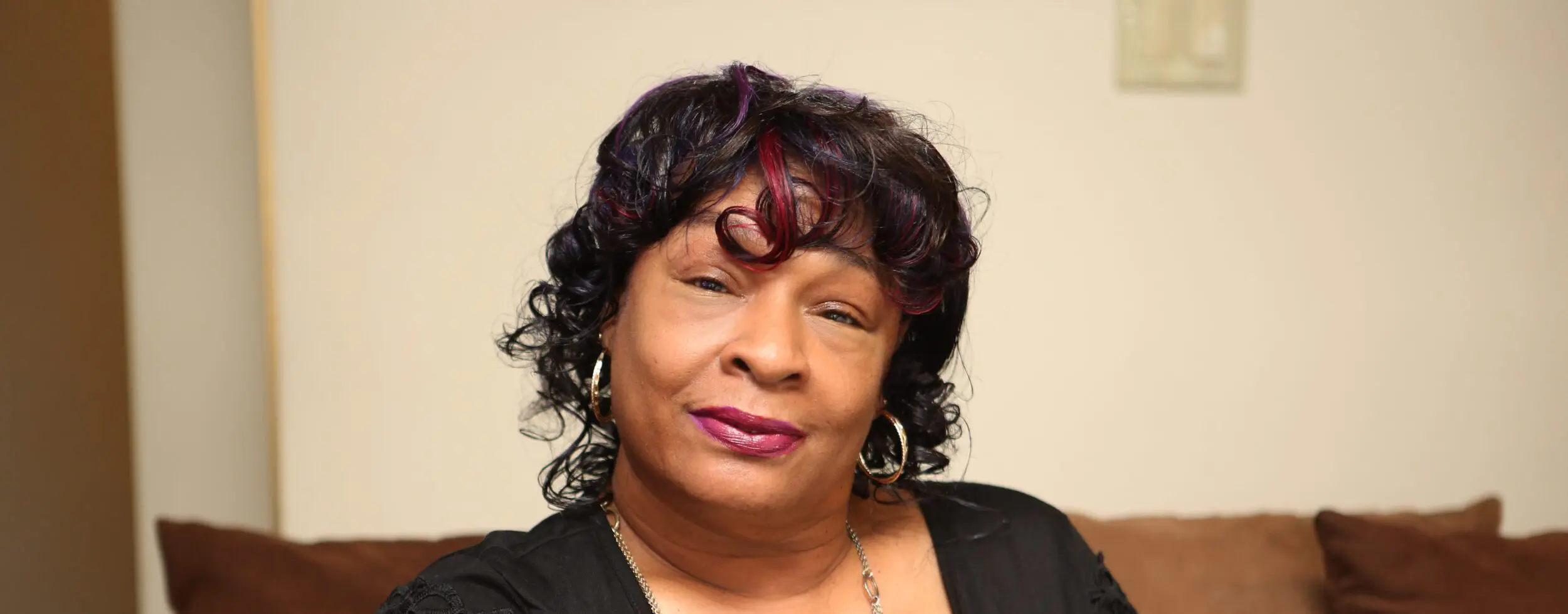 A middle-aged woman sits on a brown couch in her home.