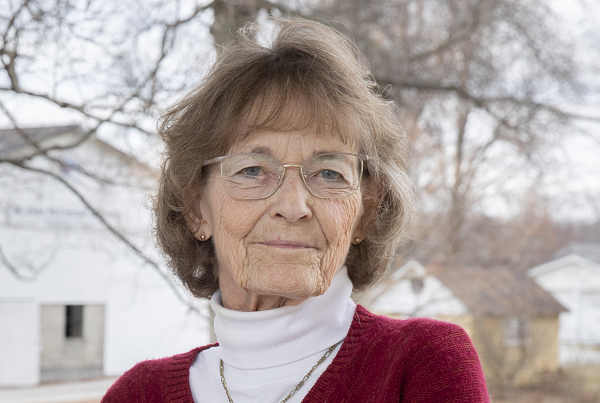 Cheryl McConnell, an elderly white woman with brunette chin-length hair, stands in front of a white barn.