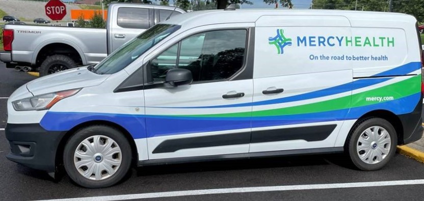 A mini van is parked in a parking lot. It is wrapped with white, blue and green Mercy Health branding.