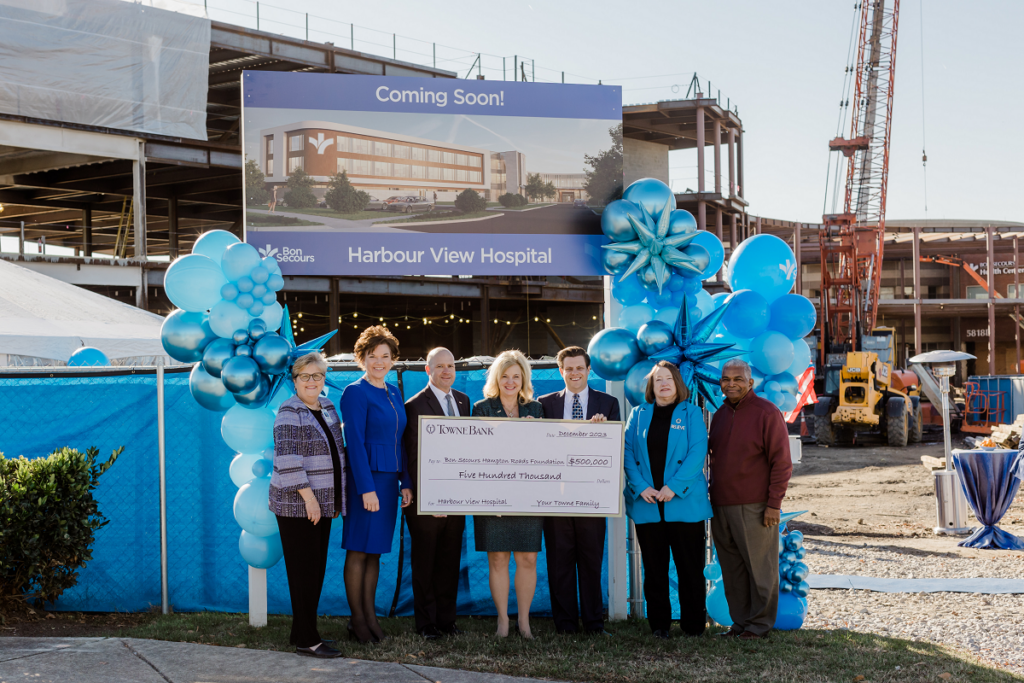 A group of business people, surrounded by balloons, exchange an XL check in front of a hospital under construction.