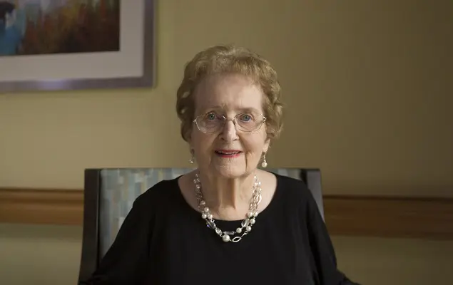 An elderly woman with short, curly hair and glasses sits indoors.