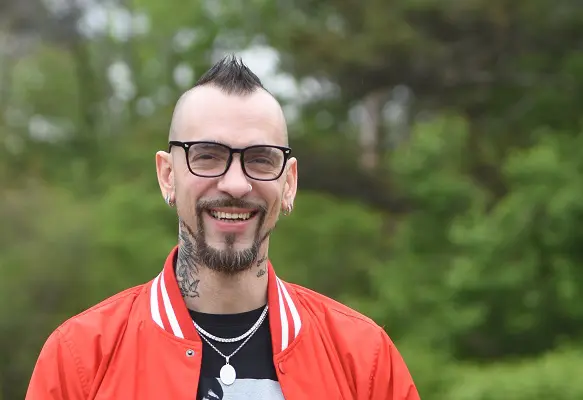 A man with a mohawk, glasses, and goatee stands outside in a park, smiling.