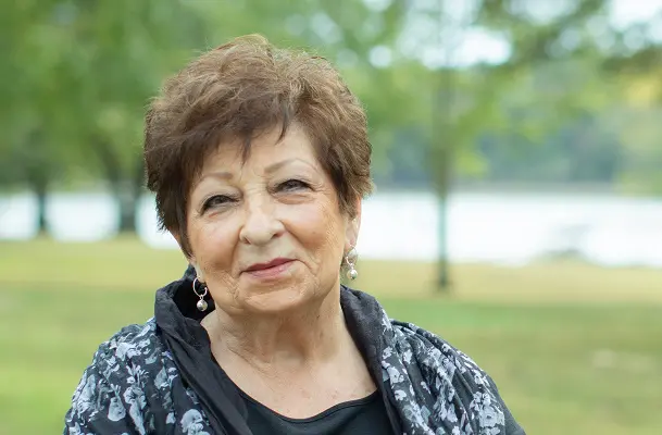 An older woman stands outside beside a lake in the summer time. She is wearing black to signify mourning.