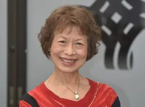 An elderly woman stands inside a gym, smiling at the camera with her head cocked happily.