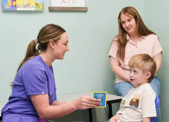 A speech therapist in purple scrubs does flashcards with a shy five-year-old boy.