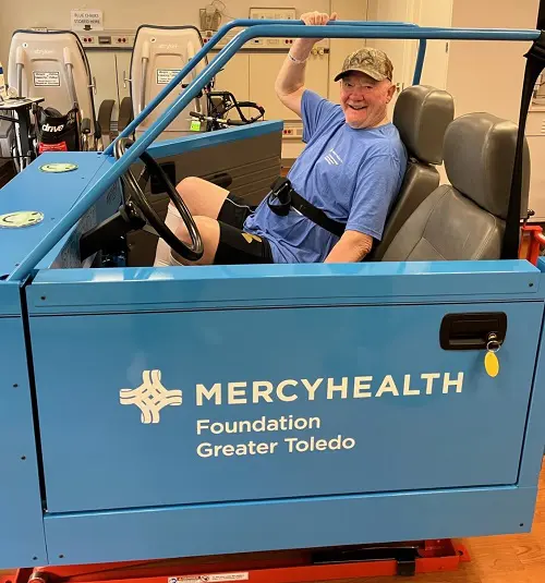 A senior man sits inside a blue car simulator at rehab. It's branded with the Mercy Health Foundation Greater Toledo logo.