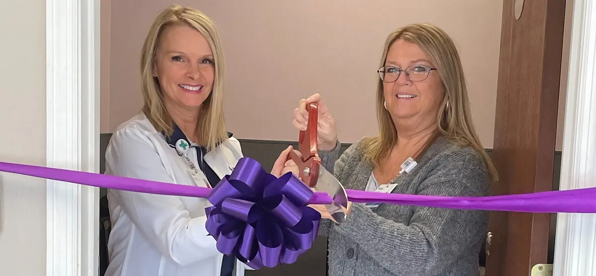 Two women use a pair of overlarge scissors to cut a purple ribbon across a room.