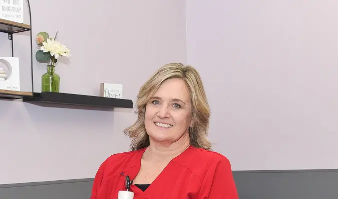 A nurse in red scrubs stands in front of a photo of trees, inside a doctor's office.