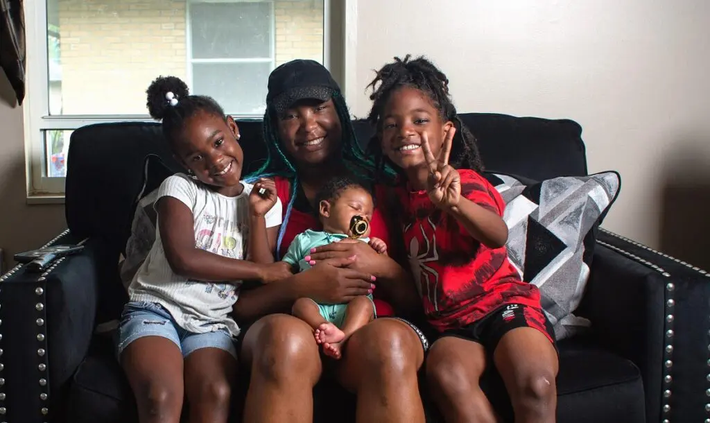 A young family sits on a couch together. Mom holds a newborn baby on her lap, while toddler-aged twins lean in from either side.