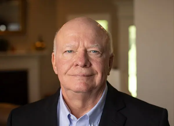 An elderly man dressed in a suit jacket smiles at the camera.