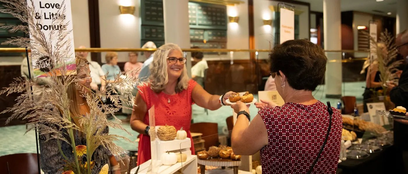 A smiling vendor hands a customer a plate of artisanal donuts at an event.