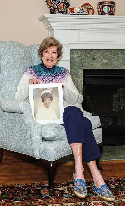 A senior woman with short, brown hair sits in a pale green armchair inside. She's holding a framed portrait of herself at a younger age, in a nurse's uniform.