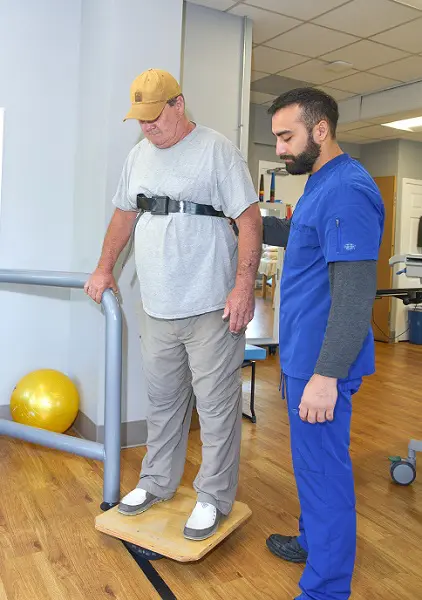 An older man in a baseball cap, t-shirt, and cargo pants stands on a balance board at a physical therapy office, under the supervision of a doctor.