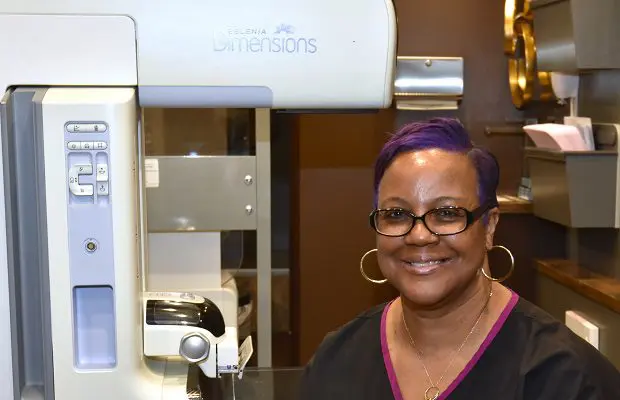 A middle-aged woman in black scrubs stands beside a mammogram machine.