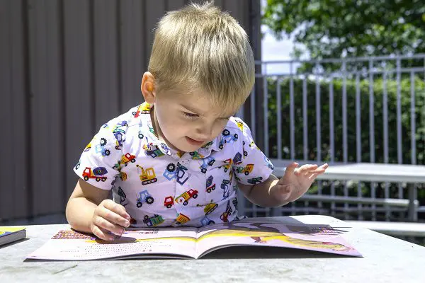 A young boy sits outside on a sunny day. He's smiling and reading at a picnic table.