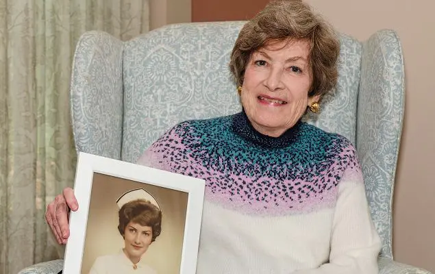 A senior woman with short, brown hair sits in a pale green armchair inside. She's holding a framed portrait of herself at a younger age, in a nursing uniform.