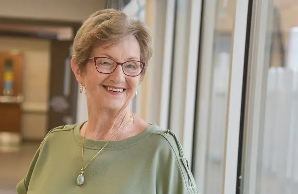 A senior woman with short, blonde hair and red-rimmed glasses smiles while standing in a well-lit hospital hallway.