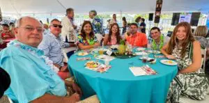 A family dressed in floral summer garments sits at an oval table outside, under a tent. The table has a bright blue table cloth on it and is laden with beverages and games.