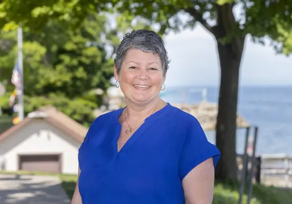 A middle-aged woman with cropped, graying hair smiles while standing outside on a summer day. Behind her is a blue, calm lake.