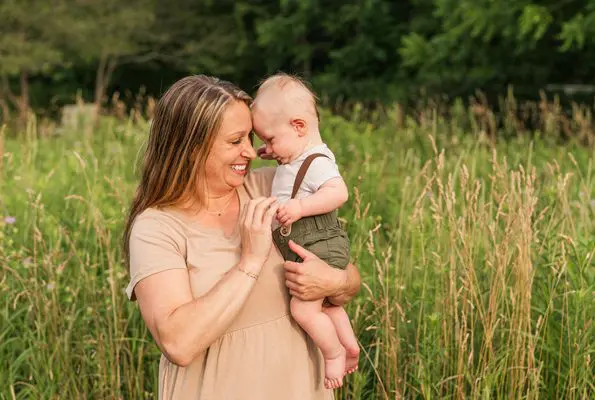A young woman stands outside, snuggling her baby on a sunny summer day.