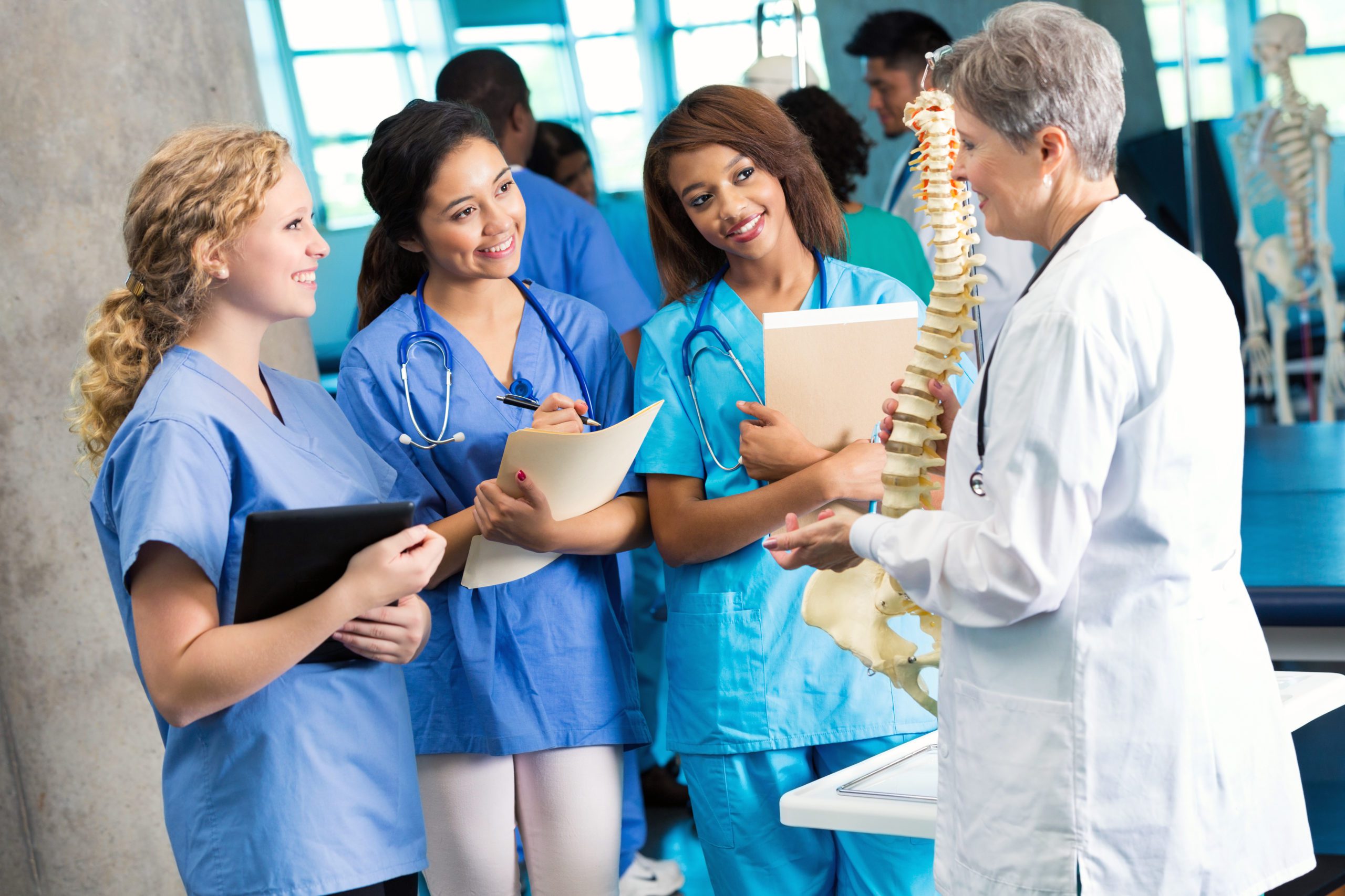 Senior Caucasian medical school professor or doctor uses a model of the human spine to teach a group of medical or nursing students. The students take notes as the professor talks about the spine. The students are wearing scrubs and the doctor is wearing a lab coat.