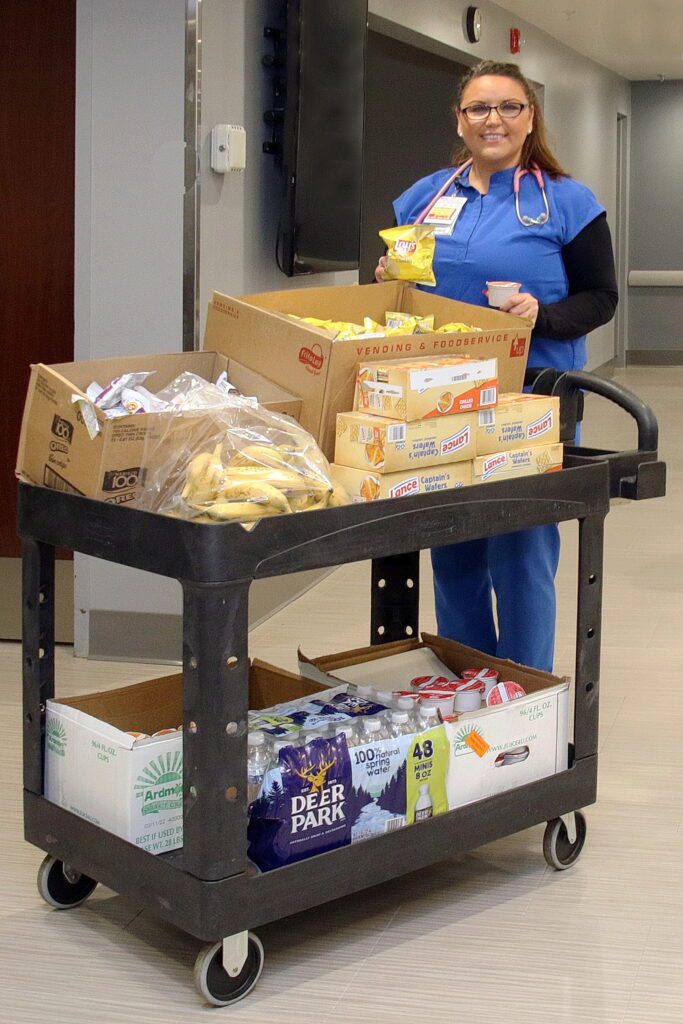 Kristy Jopek stands in a hospital hallway, wearing her scrubs, posing with a cart full of snacks.