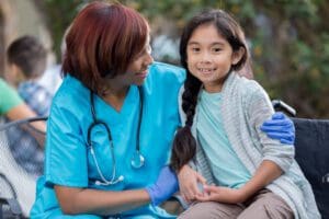 Smiling African American female nurse smiles while hugging young Asian female patient. The patient is sitting in a wheelchair. The nurse is wearing scrubs and a stethoscope.