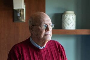 An elderly man in a red sweater stands in a hospital room, looking out the window contemplatively.