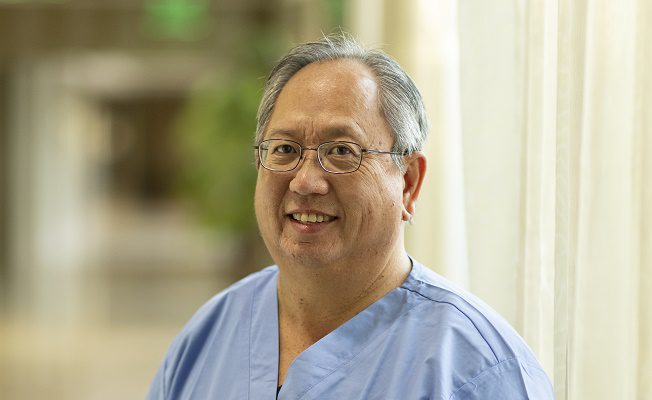 A male nurse in blue scrubs stands inside a medical facility.