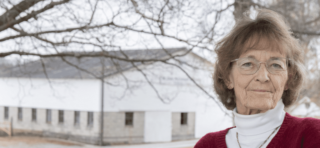 Cheryl McConnell, an elderly white woman with brunette chin-length hair, stands in front of a white barn.