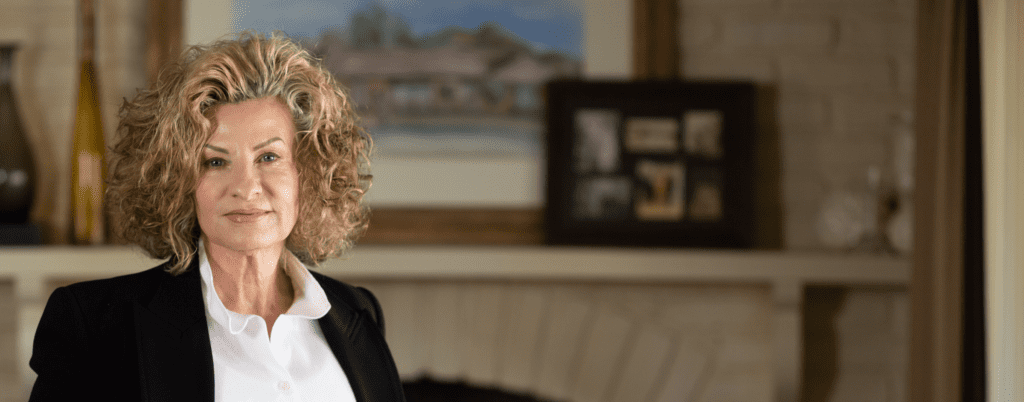 A photo of Theresa Jones, an older white woman with curly blonde hair, stands in front of a fireplace.