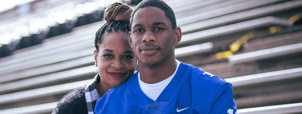 A teenage football player sits on the bleachers with his mother hugging him.