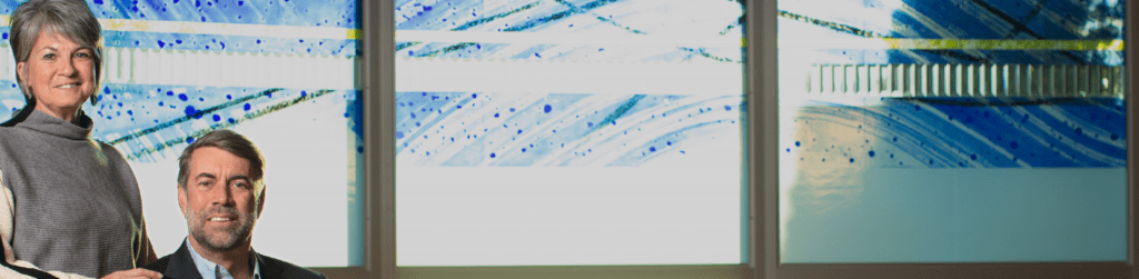 A middle-aged man and woman stand indoors in front of a stained glass window.