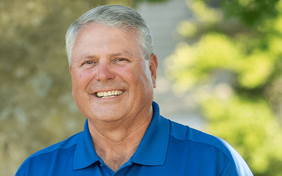 An elderly white man smiles while standing outside beneath a tree on a sunny day.