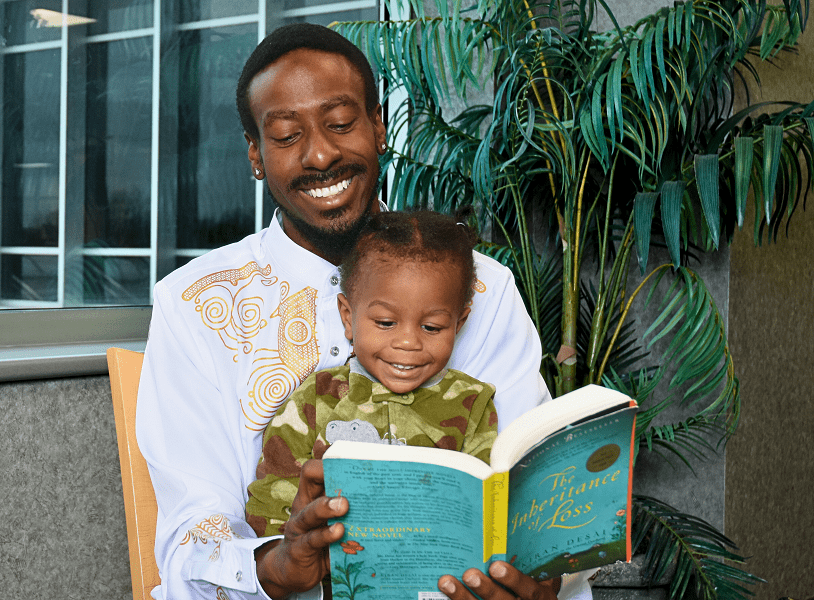 A young Black dad reads to his baby, who is sitting in his lap.