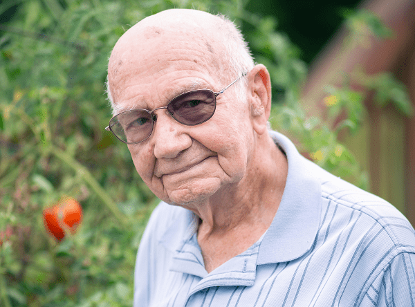 An elderly man stands outside in front of a tomato plant.