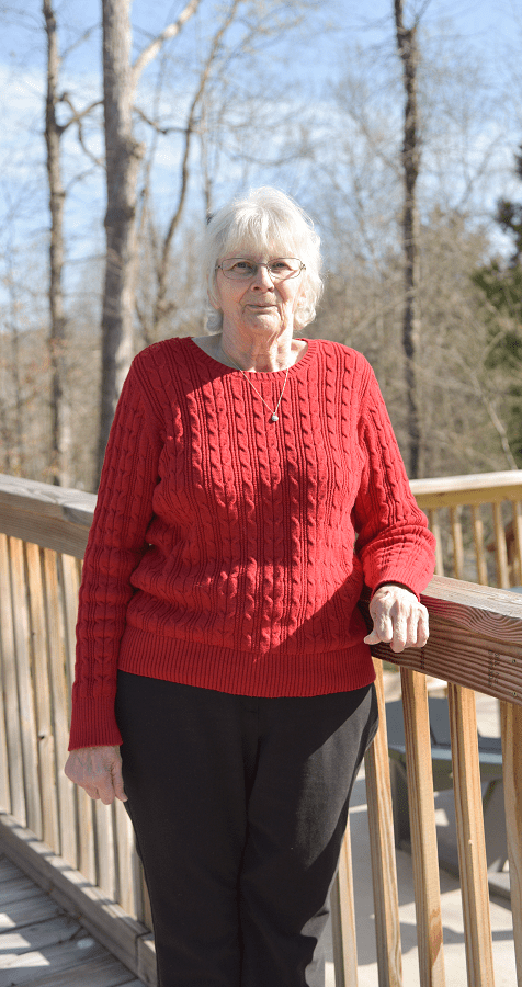 An elderly woman stands outside on a wooden deck in the sunshine.