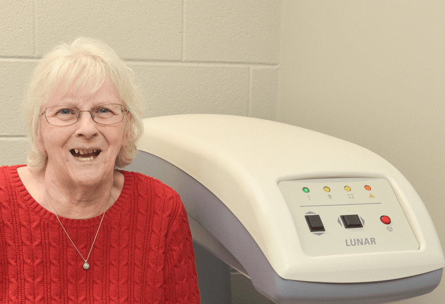 An elderly woman sits next to a DEXA machine, which is a technology that screens for osteoporosis.