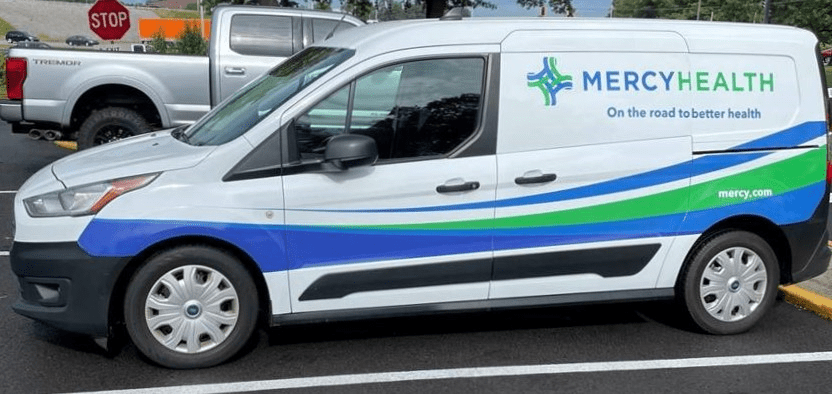A mini van is parked in a parking lot. It is wrapped with white, blue and green Mercy Health branding.
