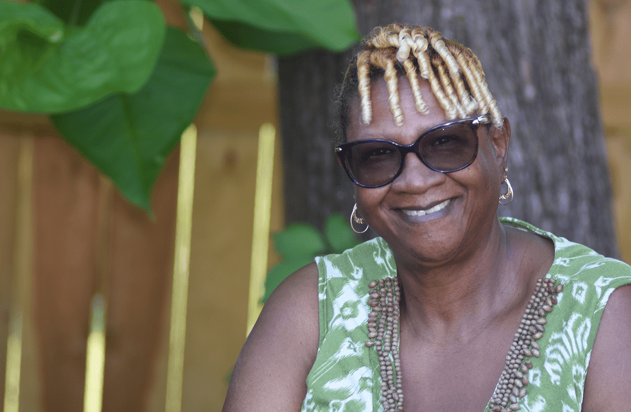 An older woman sits outside in the shade on a sunny day, smiling.