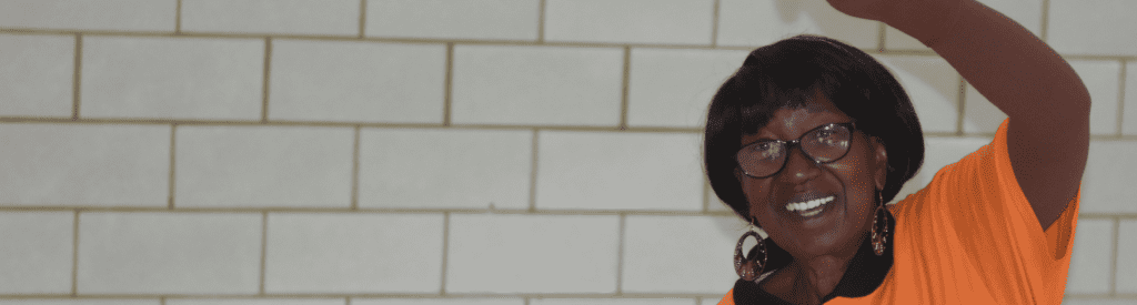 An elderly woman smiles while lifting a weight above her head in a gym.