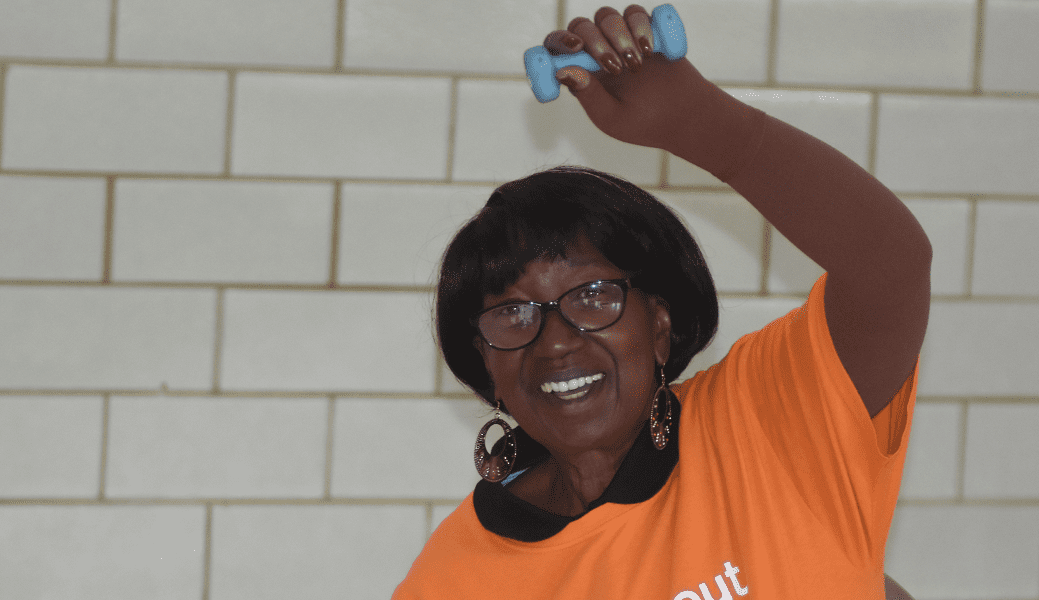 An elderly woman smiles while lifting a weight above her head in a gym.
