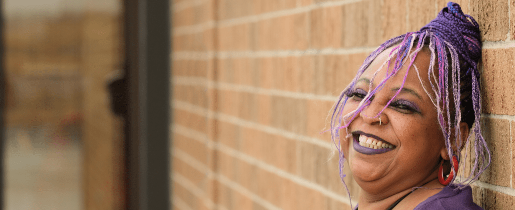 Vanessa Grimsley, a middle-aged black woman with purple braids, smiles and leans against a brick wall.