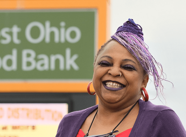 Vanessa Grimsley, a middle-aged black woman with purple braids, stands in front of the West Ohio Food Bank sign.