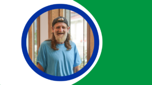 Smiling man with a long beard and hair, wearing a blue T-shirt and backward cap, standing by a window in an indoor setting.