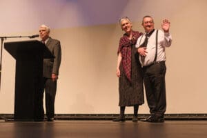 Three people stand on stage, one of whom waves to the audience