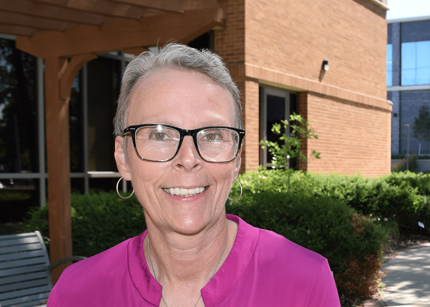 A woman with short hair and glasses smiles while sitting outside a medical building.