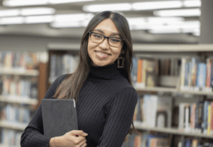 A young woman stands in a library, holding a tablet.