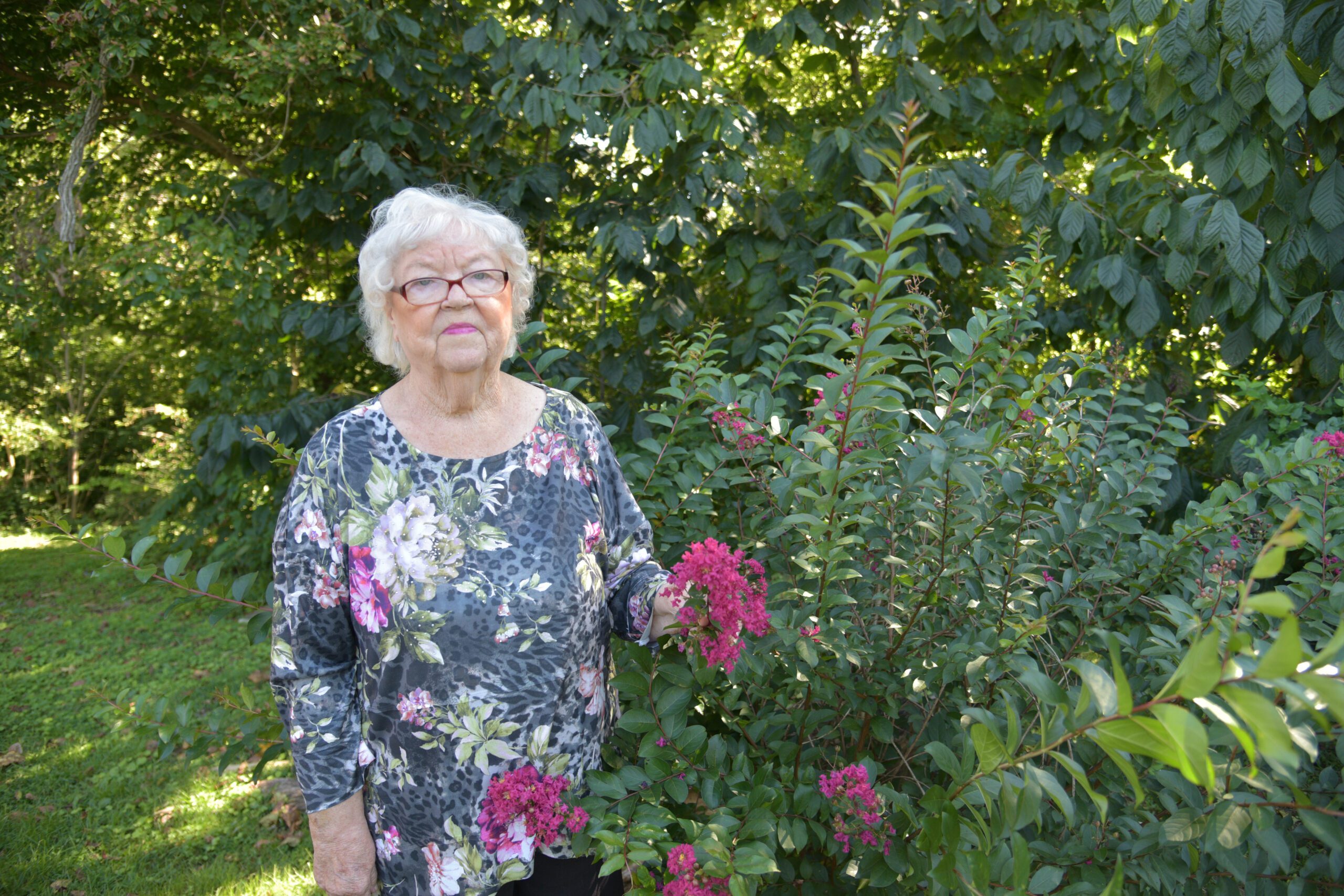 An elderly woman with white hair and glasses stands in a garden.